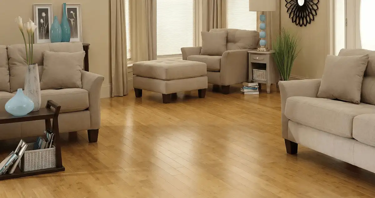 A living room with professionally installed bamboo flooring, beige sofas, a matching ottoman, a side table with a blue lamp, vases, framed pictures, and large windows with blinds and curtains in Sacramento, CA